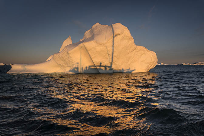 The first rays illuminate the iceberg in golden light, with the silhouette of the yacht from which it was shot, 2017