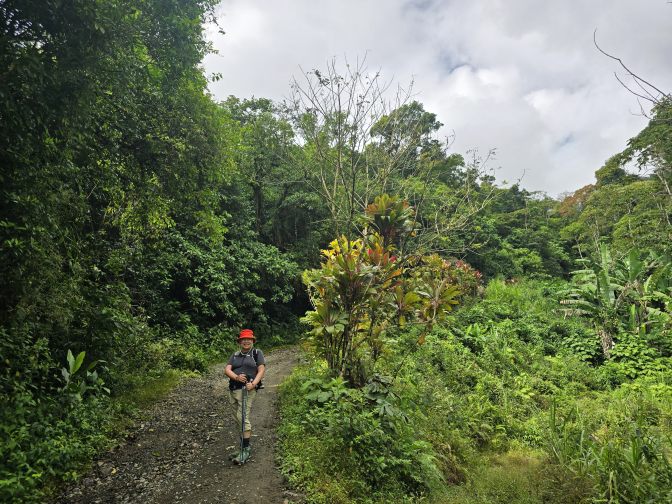 Bruna hiking along a dirt path surrounded by lush, dense rainforest vegetation, 2026